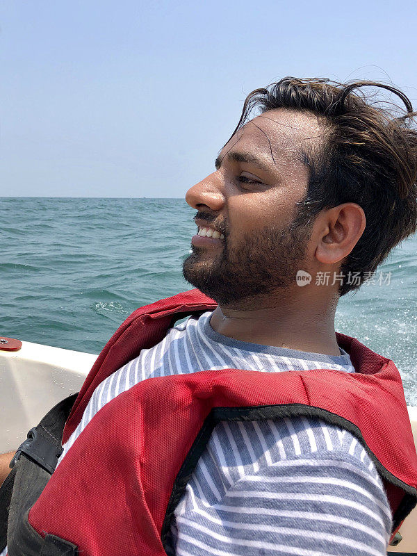 一名印度男子穿着红色救生衣乘坐快艇，在印度南部喀拉拉邦海岸线/科瓦拉姆海滩上航行，被海浪溅起水花，游船男子笑着，在船上淋湿，夏天的海边水上运动度假